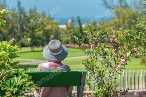 Preview: Rear view of elderly caucasian woman with hat sitting on outdoor bench in public park