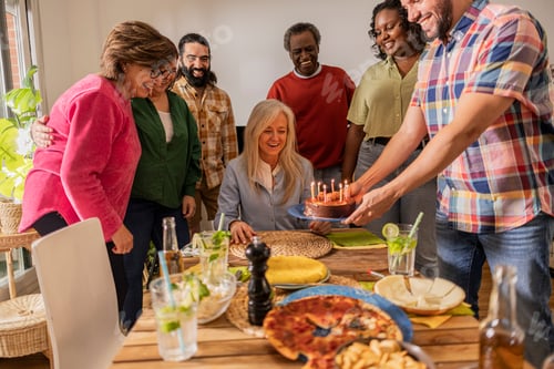 Preview: group of multiracial family and friends celebrating birthday at home with a cake with candles.