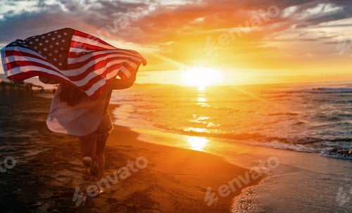 Preview: Beautiful patriotic young woman with the American flag held in her standing in the summer sunset