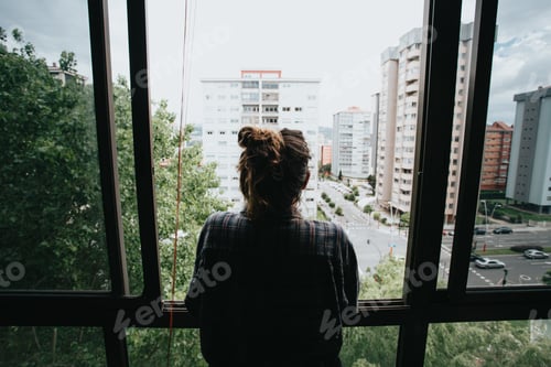 Preview: Worried woman looking from the window of her flat, mental health concept in the city