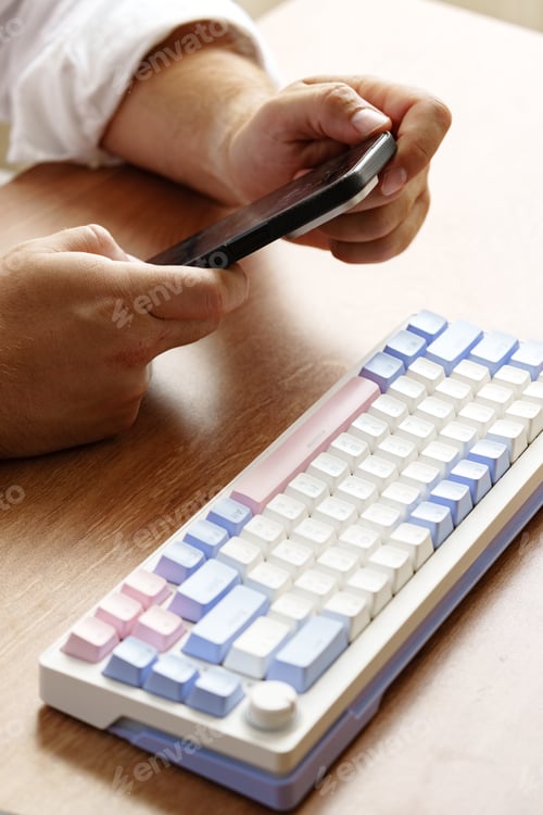 Preview: Individual using a smartphone while seated at a wooden table with a colorful keyboard