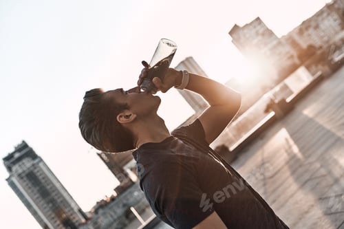 Preview: Hydrating. Young man in sports clothing drinking water while standing outside