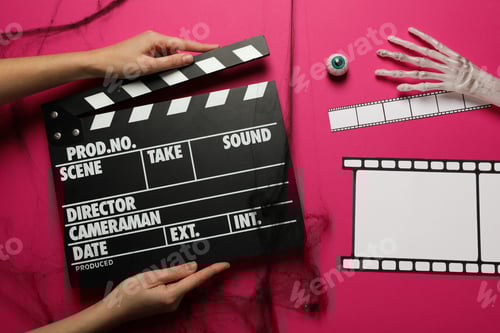 Preview: Clapperboard in hand, film and Halloween decorations on pink background, top view
