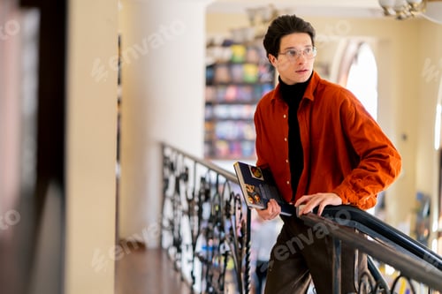 Preview: portrait of a young guy in glasses standing with a book in his hands in library