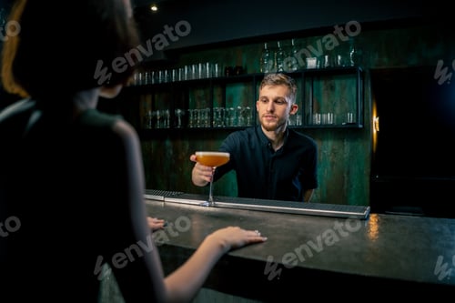 Preview: in a night bar young bartender with a beard in a black shirt is preparing a cocktail for a client at