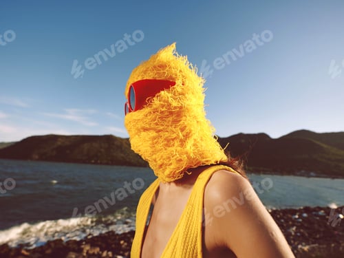Preview: Bird mask woman by ocean in front of mountains a woman wearing a yellow bird mask stands near the