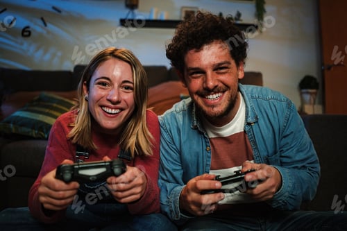 Preview: Young couple having fun plaing video games with a console together sitting at couch at home. Two