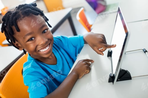 Preview: Cheerful African American boy sitting at desk in classroom, pointing at white tablet on black stand