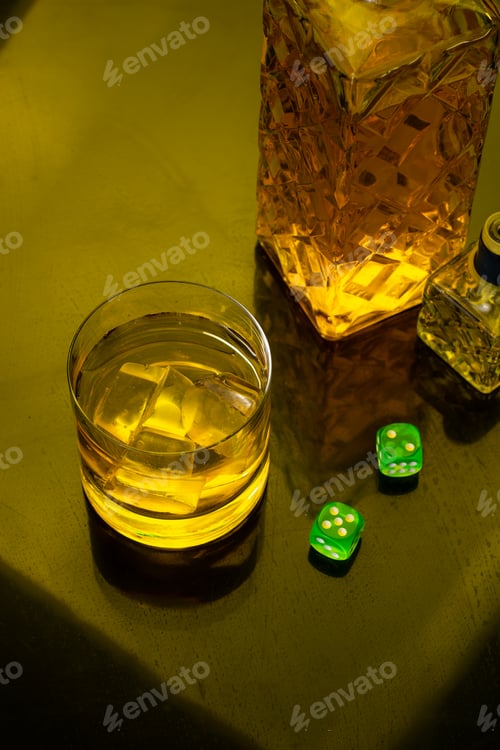 Preview: Top view of a glass of Whiskey with ice next to a bottle, green dice on glass table