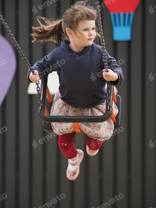 Preview: Little cute stylish girl in brightly colored swings on a swing at the playground on a summer day
