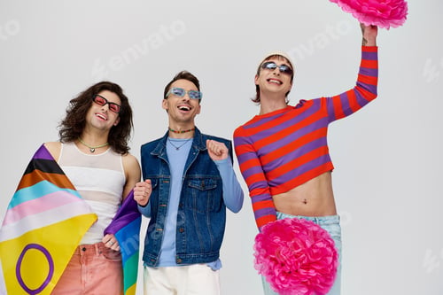 Preview: three cheerful stylish gay men in bold clothes with sunglasses posing with pom poms and rainbow flag