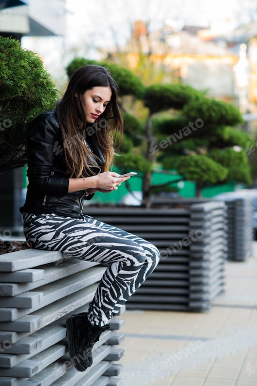 Preview: Beautiful young dark-haired woman is texting on her smartphone while sitting the bench in city park