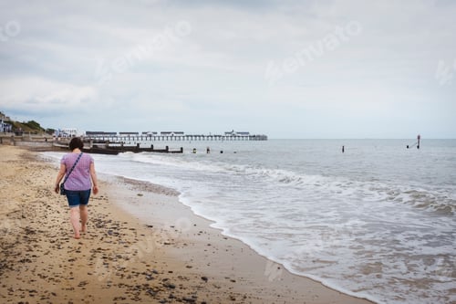 Preview: Rear view of woman walking barefoot along a sandy beach by the ocean.
