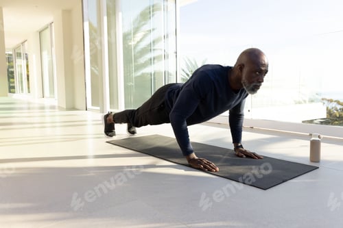 Preview: Senior African American man holding plank on mat in sunroom wearing navy top, tracker, water bottle