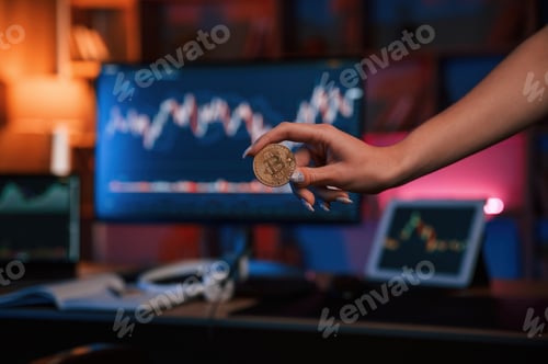 Preview: Close up view of woman's hand that holding bitcoin against computer display and tablet with graphs