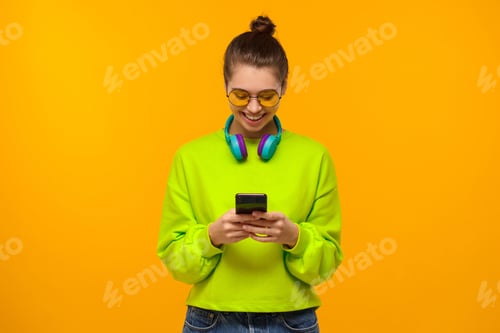 Preview: Teen girl wearing headphones around neck, looking at screen of phone with smile