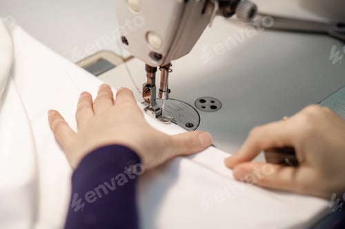Preview: Hands of a dressmaker sewing clothes from white fabric on a sewing machine, close-up.