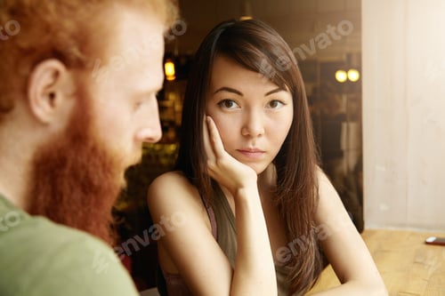 Preview: Multiethnic couple. Selective focus. Asian girl with worried look, resting elbow on table, sitting i