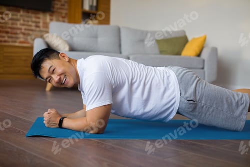 Preview: Happy asian mature man doing fitness training on sports mat at home, standing in elbow plank and