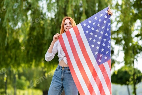 Preview: Happy teenage girl posing with USA national flag standing outdoors in summer park.