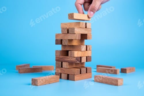 Preview: Hand placing wooden block on unstable tower on blue background