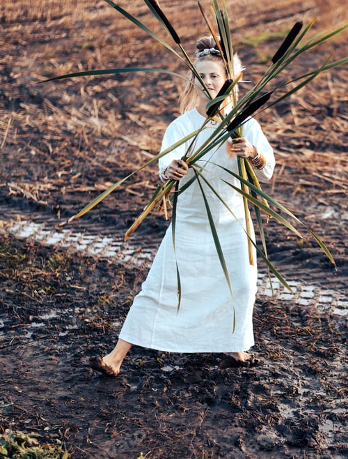 Preview: Beautiful woman in a dress dances barefoot on a muddy field, holding reeds in her hands