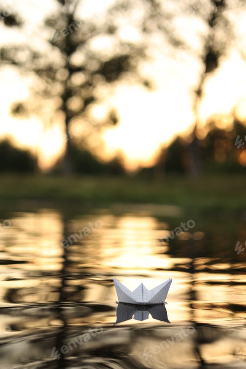 Preview: A paper boat is floating on the waves in the water at a beautiful sunset. Origami ship Sailing. T