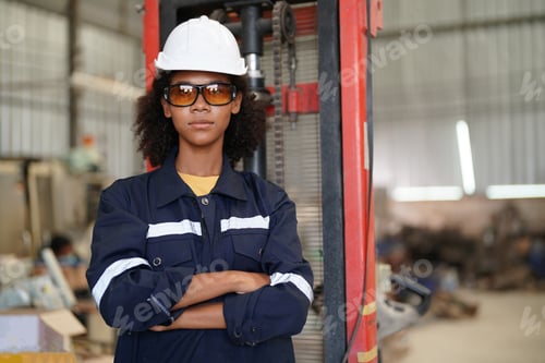 Preview: Confident black female engineer at a manufacturing plant facing camera smiling with arms crossed wea