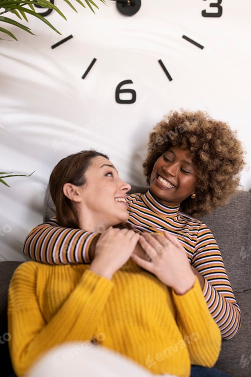 Preview: Multiracial lesbian couple hugging and laughing and relaxing in a living room. Smiling gay females