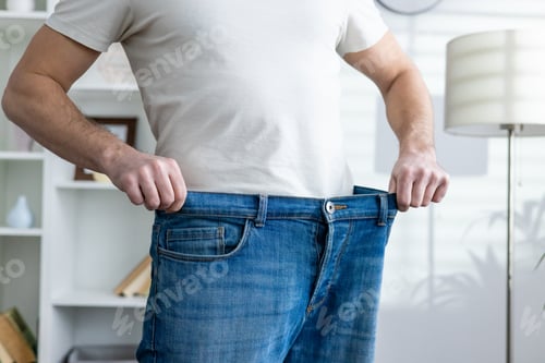 Preview: Man showing weight loss by wearing loose jeans in a bright room