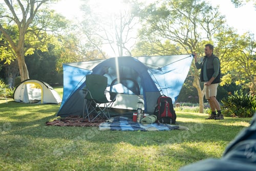 Preview: Man pitching blue dome tent on sunlit clearing with picnic blankets, camping chair and backpack
