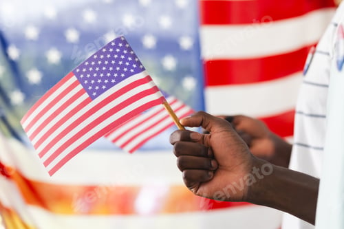 Preview: African American man's hand holding a small American flag, with a larger flag in the background