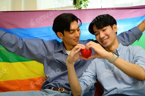 Preview: Smiling young men couple holding a red heart and sitting under rainbow pride flag
