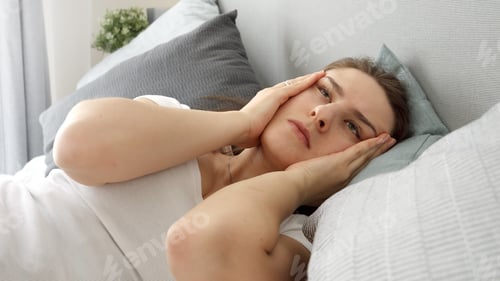 Preview: Portrait of young woman feeling pain in head holding hands on temples while lying in bed