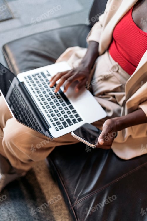Vista previa: Vista superior de una mujer irreconocible usando su portátil y su teléfono sentada en un sofá
