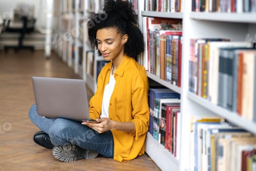 Preview: Modern focused pretty mixed race black female student with curly hair, sitting on a floor in library