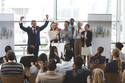Preview: Businessman standing with arms up to victory at podium with colleagues at business seminar in office