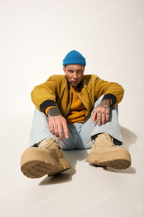 Preview: Stylish young man with tattoos poses in casual attire against a simple backdrop