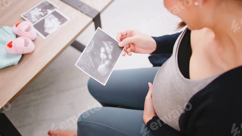 Preview: Happy Asian pregnant woman watching ultrasound image on sofa.