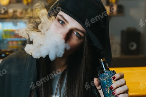 Preview: Young pretty woman in cap smoke an electronic cigarette at the vape shop. Hip-hop style. Closeup