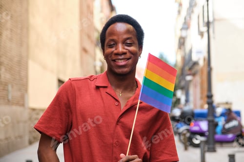 Preview: Gay black man in a red shirt is holding a rainbow flag and smiling at Pride Day.
