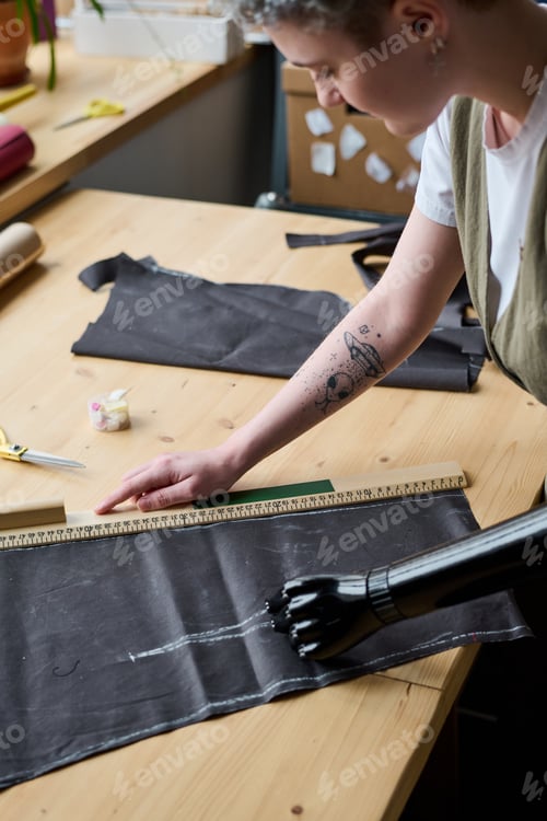 Preview: Female fashion designer bending over table with textile workpiece and ruler