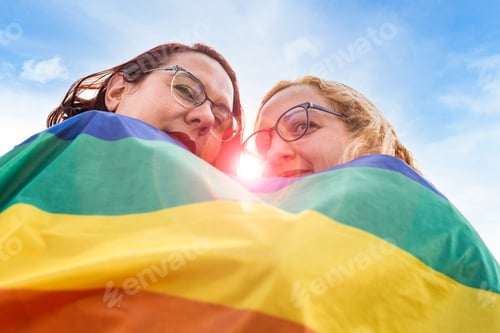 Preview: Low angle view of lesbian couple under LGBT rainbow flag while looking at camera.