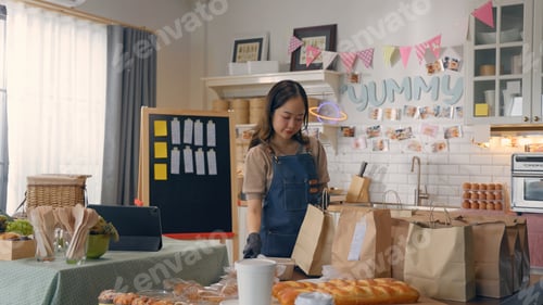 Preview: Woman preparing baked goods in cozy kitchen setup, surrounded by bread and paper bags, ready for del