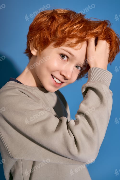 Preview: Stylish little boy smiles while playing with his hair in front of a vibrant blue backdrop