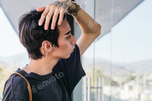 Preview: Side view of stylish homosexual man touching hair and looking in glass wall of building in street