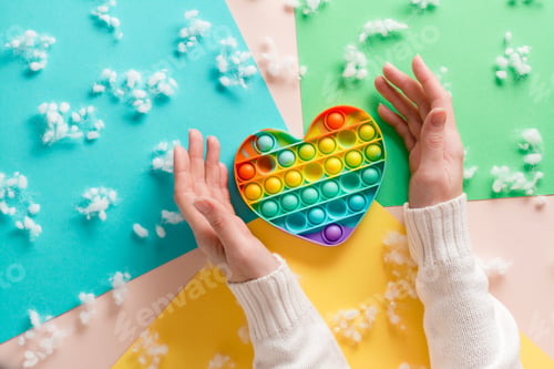 Preview: Female hands surround a silicone rainbow antistress toy in the shape of a heart