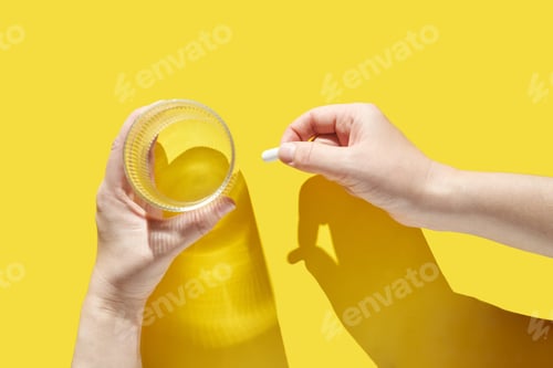 Preview: Caucasian female hands holding water glass and medicine pill on vibrant yellow