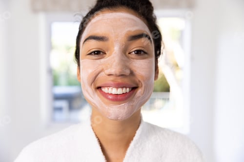 Preview: Portrait of happy biracial woman in bathrobe with face mask in sunny bathroom