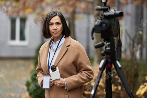 Preview: Multiracial female journalist reporting outside in a beige coat during autumn